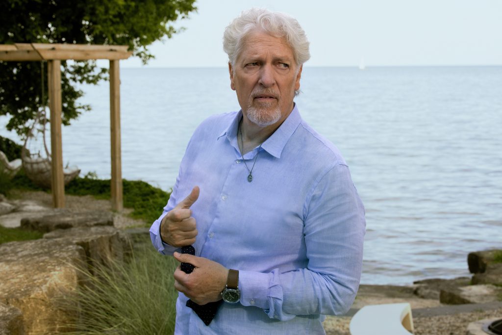 "Older man with white hair and beard stands outdoors by a lakeshore, wearing a light blue shirt and holding a necktie. Natural background with rocks, trees, and water on a clear day."